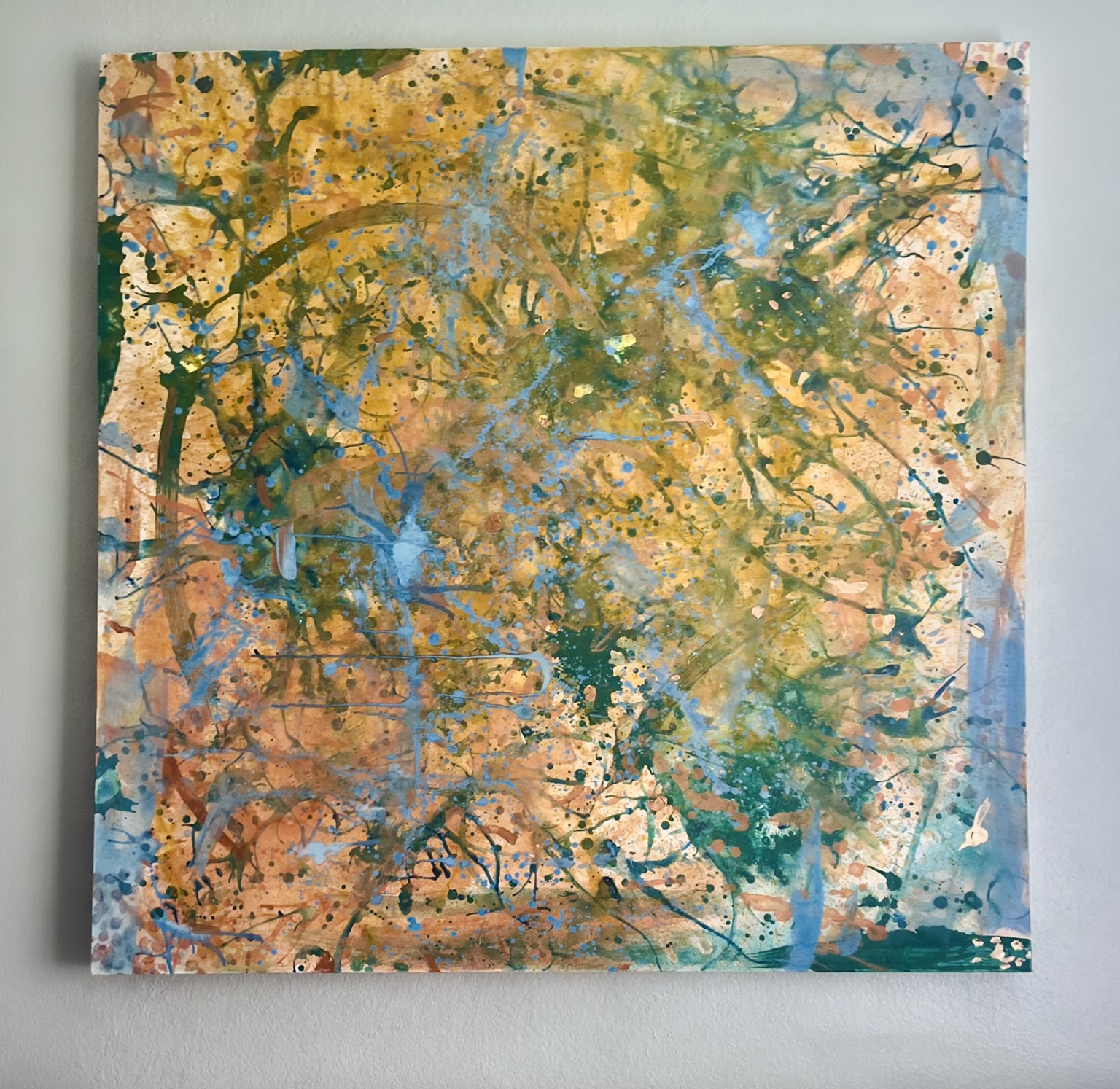 Large abstract painting displayed on a white wall, with layers of ochre, peach, teal, green, and blue splatters